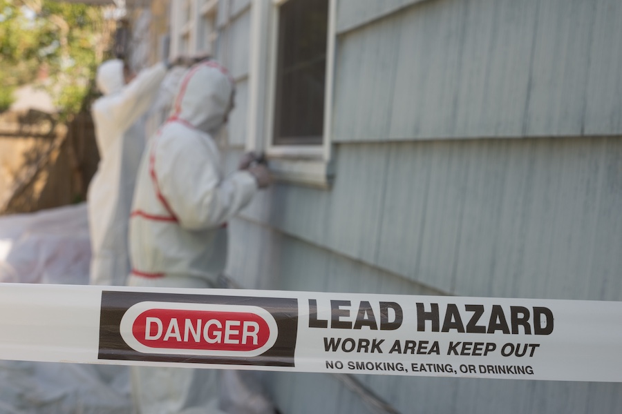 Hazard tape reads 'DANGER LEAD HAZARD WORK AREA KEEP OUT' in foreground as two workers in protective hazmat suits work near a house window in the background.