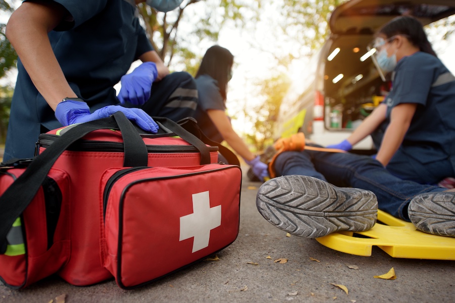 Emergency responders treat a patient on a yellow spine board beside an ambulance; red first-aid bag in foreground with a white cross.