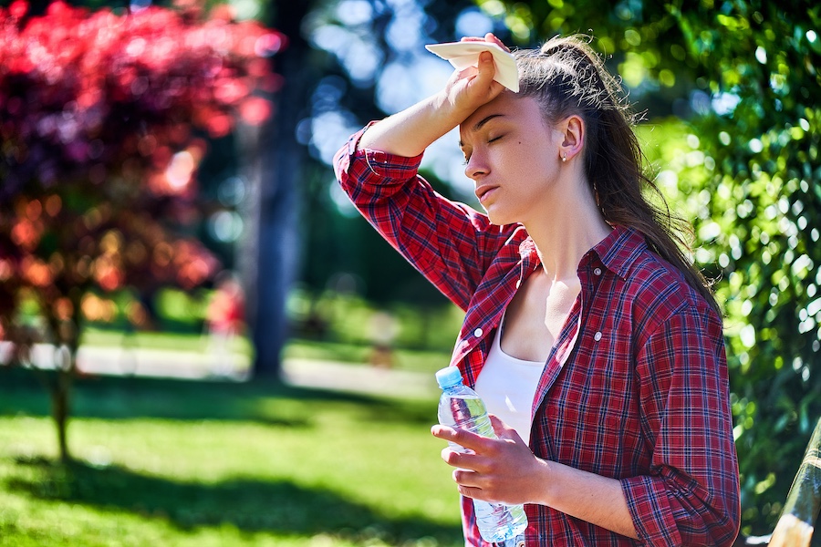 Woman in a red plaid shirt wipes sweat from her forehead with a tissue in a sunny park, holding a water bottle in the other hand.