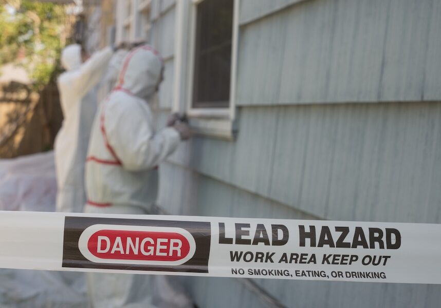Hazard tape reads 'DANGER LEAD HAZARD WORK AREA KEEP OUT' in foreground as two workers in protective hazmat suits work near a house window in the background.