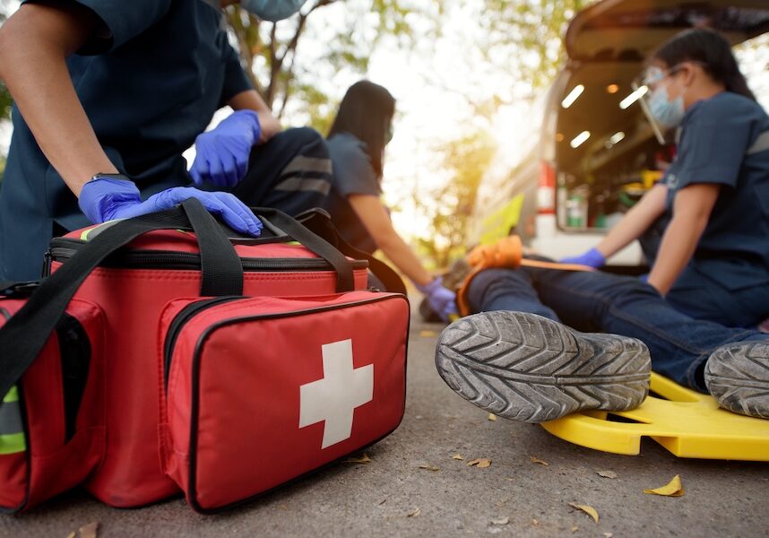 Emergency responders treat a patient on a yellow spine board beside an ambulance; red first-aid bag in foreground with a white cross.