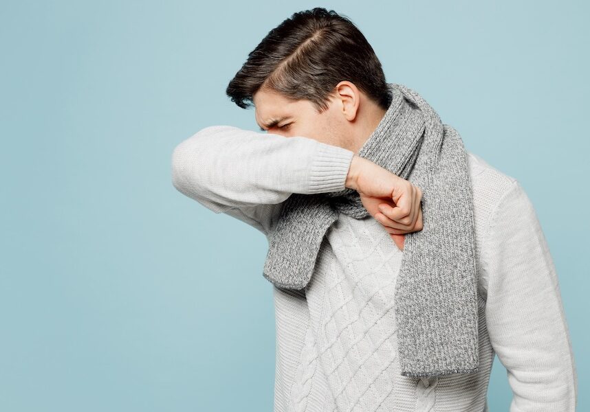 Young ill sick man wear gray sweater scarf cough cover mouth with hand sleeve isolated on plain blue background studio portrait. Healthy lifestyle disease virus treatment cold season recovery concept