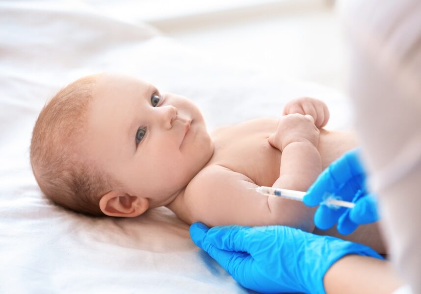Baby lying on a bed as a clinician in blue gloves administers a vaccine in the upper arm.