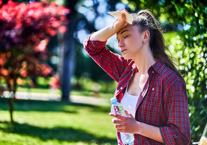 Woman in a red plaid shirt wipes sweat from her forehead with a tissue in a sunny park, holding a water bottle in the other hand.